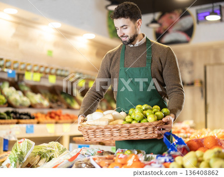 Man employees in uniform carries wicker basket with various vegetables and fruits in grocery shop 136408862