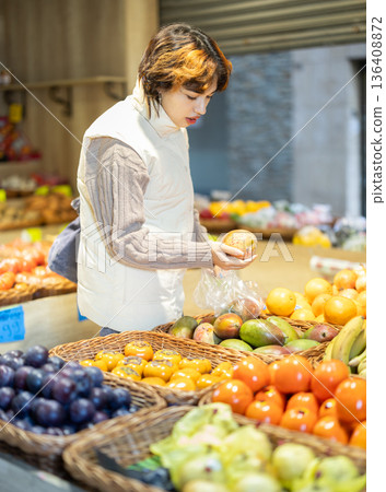 Woman chooses yellow mango for purchase in greengrocer shop Woman chooses yellow mango for purchase in greengrocer shop 136408872