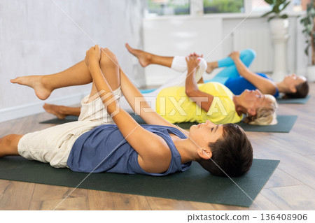 Preteen boy practicing knee to chest pose of yoga in light fitness room 136408906