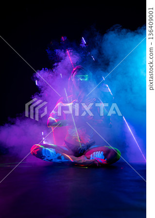 Woman in UV body paint and cyber glasses sits in a meditative pose amidst purple smoke and laser beams. Woman in UV body paint and cyber glasses sits in a meditative pose amidst purple smoke and laser beams. 136409001