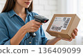 Woman scanning a shipping label on a cardboard box. E-commerce order fulfillment with a barcode scanner 136409099