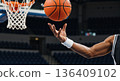 Close-up of a basketball player's muscular arm tossing a ball. Athlete training in an arena with a hoop in the background. Professional sports action 136409102