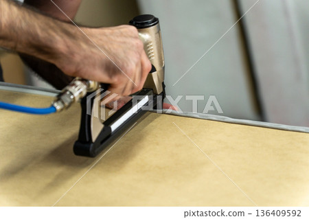 Close-up of upholsterer using a pneumatic staple gun to attach fabric to a wooden furniture frame. 136409592