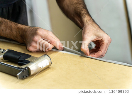 Close-up of upholsterer hands folding fabric edge on a wooden furniture frame 136409593