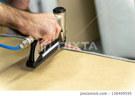 Close-up of upholsterer using a pneumatic staple gun to attach fabric to a wooden furniture frame. 136409595