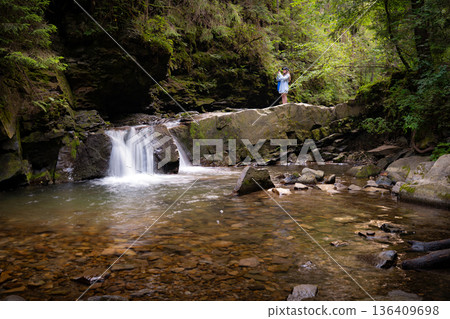 Woman Taking Photos on Stone Ledge Above Forest Waterfall 136409698