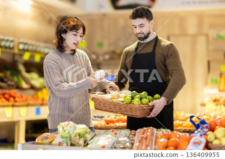 Girl customer in vegetable store examines radish, male seller advises and help with choice Girl customer in vegetable store examines radish, male seller advises and help with choice 136409955