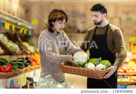 Girl customer in vegetable store examines cauliflower, male seller advises and help with choice Girl customer in vegetable store examines cauliflower, male seller advises and help with choice 136409966
