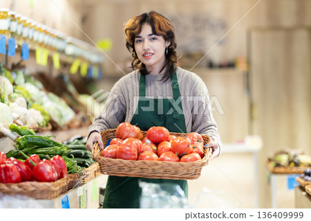 Girl employees in uniform carries a wicker basket with tomatoes in grocery shop 136409999