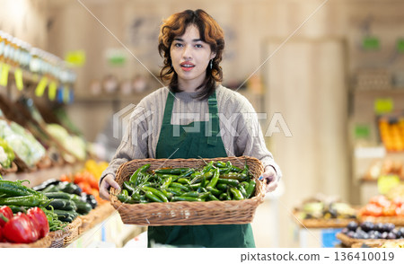 Woman employees in uniform carries wicker basket with green peppers in grocery shop Woman employees in uniform carries wicker basket with green peppers in grocery shop 136410019
