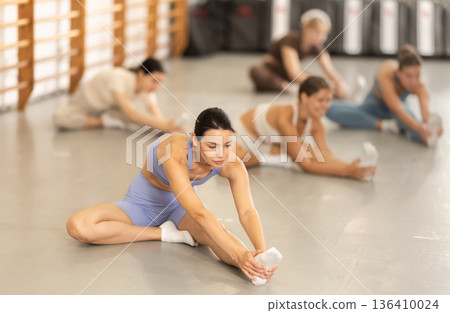 Group of girls in gym do stretching, sitting on floor and bending towards legs. Group of girls in gym do stretching, sitting on floor and bending towards legs. 136410024