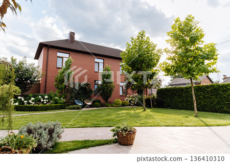 Brick family house with landscaped backyard, green lawn, trees and patio in sunlight Brick family house with landscaped backyard, green lawn, trees and patio in sunlight 136410310