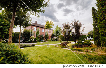 Spacious landscaped backyard with green lawn, trees, patio paths and brick house under cloudy sky Spacious landscaped backyard with green lawn, trees, patio paths and brick house under cloudy sky 136410314