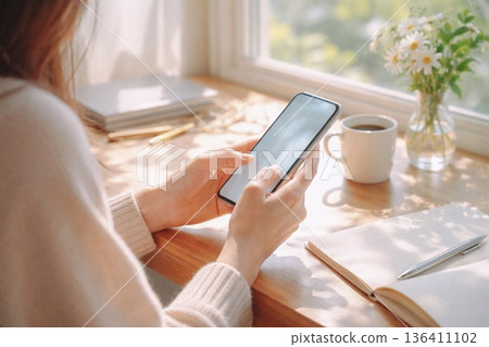 清晨的陽光灑進窗邊，一隻女人的手放在桌上，手裡拿著一支智慧型手機。 136411102