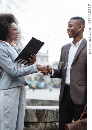 Two professionals, a man and a woman, shake hands outdoors while discussing business matters and reviewing documents Two professionals, a man and a woman, shake hands outdoors while discussing business matters and reviewing documents 136412227