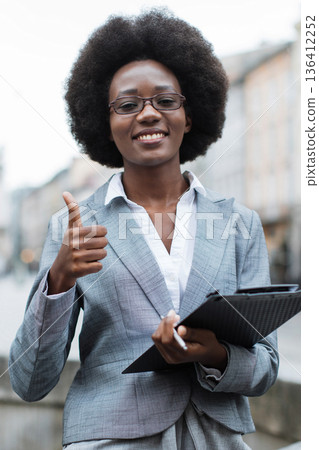A smiling Black woman in a grey suit and glasses holds a clipboard and gives a thumbs-up gesture, conveying approval and success outdoors A smiling Black woman in a grey suit and glasses holds a clipboard and gives a thumbs-up gesture, conveying approval and success outdoors 136412252