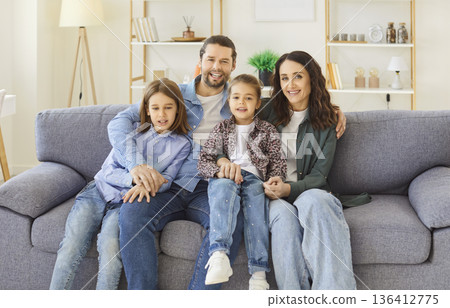 Happy parents sitting on sofa with two kids, hugging and smiling while looking at camera at home. Happy parents sitting on sofa with two kids, hugging and smiling while looking at camera at home. 136412775
