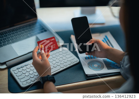 Person Holding Credit Card and Smartphone While Sitting at Desk with Laptop and Graphs on Papers, Financial Transactions Concept in Modern Office Environment Person Holding Credit Card and Smartphone While Sitting at Desk with Laptop and Graphs on Papers, Financial Transactions Concept in Modern Office Environment 136412923