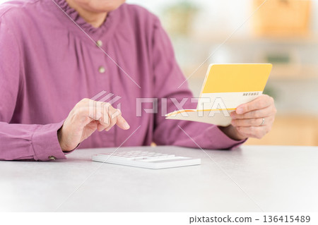 一位老年婦女一邊查看儲蓄存摺(退休金、生活開支、退休金),一邊使用計算機進行計算。 一位老年婦女一邊查看儲蓄存摺(退休金、生活開支、退休金),一邊使用計算機進行計算。 136415489