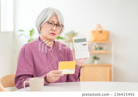 一位老年婦女一邊查看儲蓄存摺(退休金、生活開支、退休金),一邊使用計算機進行計算。 一位老年婦女一邊查看儲蓄存摺(退休金、生活開支、退休金),一邊使用計算機進行計算。 136415491