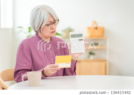 一位老年婦女一邊查看儲蓄存摺(退休金、生活開支、退休金),一邊使用計算機進行計算。 一位老年婦女一邊查看儲蓄存摺(退休金、生活開支、退休金),一邊使用計算機進行計算。 136415492