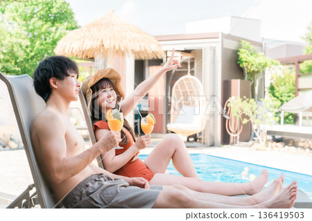 旅行中,一對情侶穿著泳裝,坐在度假泳池邊的躺椅上。 旅行中,一對情侶穿著泳裝,坐在度假泳池邊的躺椅上。 136419503