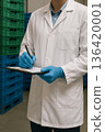 Food safety inspector in white coat and blue gloves writing notes on clipboard during audit and inspection in a food storage facility with stacked crates. 136420001