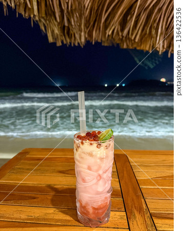Pink tropical cocktail on wooden table by night sea Pink tropical cocktail on wooden table by night sea 136422536
