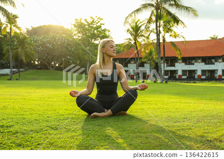 A woman practices yoga in a green field during sunset A woman practices yoga in a green field during sunset 136422615