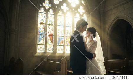 一對身著結婚禮服的日本新郎新娘在教堂裡擁抱，教堂的彩色玻璃窗照亮了整個畫面。 136423411