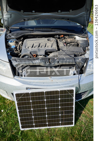 Solar panel in front of car with hood open, symbolizing transition from traditional internal combustion engines to electric cars. Charging cable connects solar panel to vehicle, renewable energy usage Solar panel in front of car with hood open, symbolizing transition from traditional internal combustion engines to electric cars. Charging cable connects solar panel to vehicle, renewable energy usage 136426663