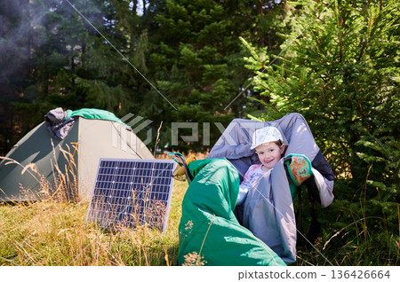 Young child playing near tourist tent with photovoltaic solar panel on summer. On background of dense forest. Integration of renewable energy in outdoor camping activities. Young child playing near tourist tent with photovoltaic solar panel on summer. On background of dense forest. Integration of renewable energy in outdoor camping activities. 136426664