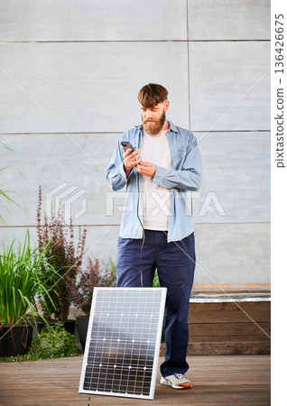 Bearded man connecting smartphone to photovoltaic solar panel. Integration of sustainable renewable energy into everyday life, demonstrating practical use of solar power for charging devices. Bearded man connecting smartphone to photovoltaic solar panel. Integration of sustainable renewable energy into everyday life, demonstrating practical use of solar power for charging devices. 136426675