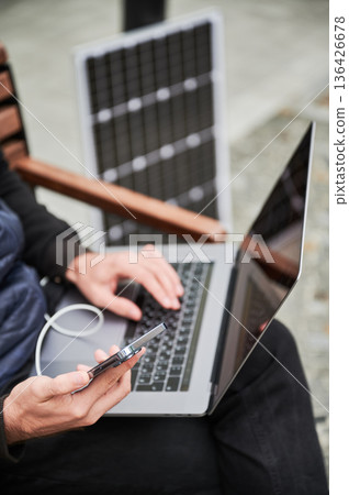 Cropped view of man sits on bench, working on laptop and smartphone, both connected to solar panel. Urban setting and casual attire highlight modern, sustainable, mobile workspace. 136426678