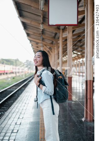 A young woman with a backpack smiles on a train platform, embodying the excitement of travel and exploration in an urban environment. A young woman with a backpack smiles on a train platform, embodying the excitement of travel and exploration in an urban environment. 136427403