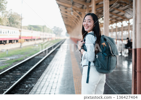 Smiling woman with backpack standing on a train station platform, ready for travel. Bright, sunny day with trains in the background. 136427404