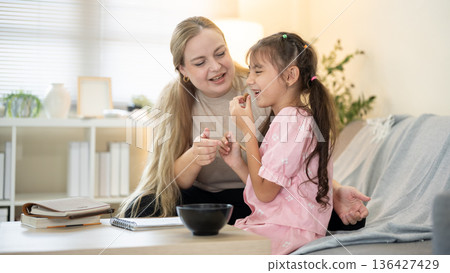 Smiling caucasian mother looking at kid daughter eating cookie while sitting on sofa in living room. Smiling caucasian mother looking at kid daughter eating cookie while sitting on sofa in living room. 136427429