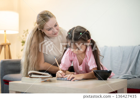 Caucasian mother looking at daughter holds pencil doing homework on table aside sofa in living room. 136427435