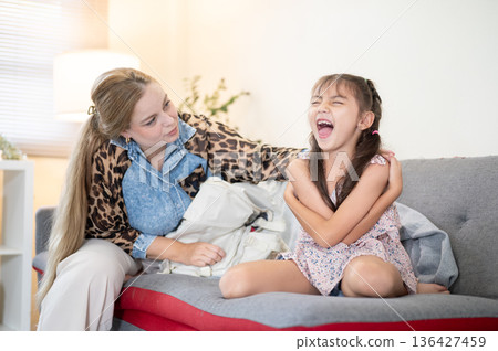 Caucasian whites mother looking at laughing asian kid daughter while sitting on sofa in living room. 136427459
