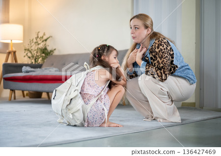 Caucasian whites mother talking to asian kid daughter with school bag on carpet in living room. 136427469