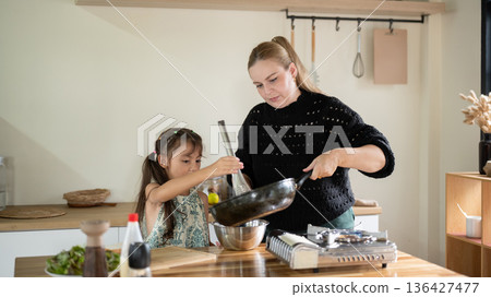 Caucasian whites mother holding pan off stove cooking with kid daughter standing at kitchen counter. Caucasian whites mother holding pan off stove cooking with kid daughter standing at kitchen counter. 136427477