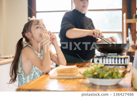 Asian daughter eating vegetable while white mother frying cooking in pan on stove at kitchen counter Asian daughter eating vegetable while white mother frying cooking in pan on stove at kitchen counter 136427486