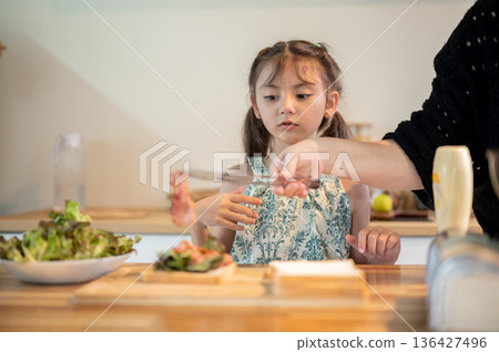 Asian daughter looking at mother putting bacon over bread making sandwich at kitchen cooking counter 136427496
