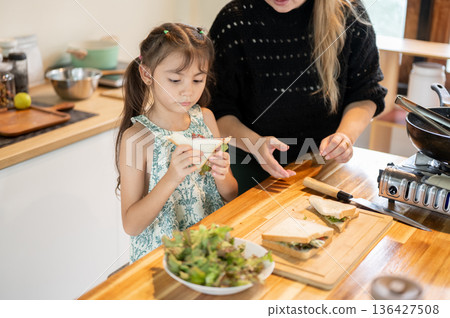Asian daughter holding a half cuts sandwich while standing aside mother at kitchen cooking counter. 136427508