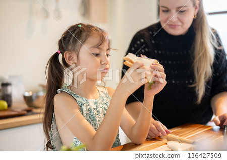 Asian daughter holding a half cuts sandwich standing with white mother at kitchen cooking counter. Asian daughter holding a half cuts sandwich standing with white mother at kitchen cooking counter. 136427509