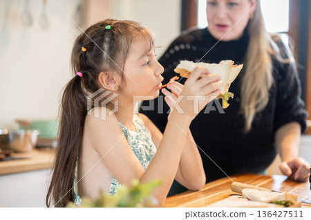 Asian daughter enjoy eating a sandwich while standing with white mother at kitchen cooking counter. Asian daughter enjoy eating a sandwich while standing with white mother at kitchen cooking counter. 136427511