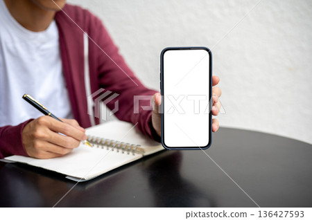 A man or student showing white screen smartphone while writing in notebook on black table in a cafe. A man or student showing white screen smartphone while writing in notebook on black table in a cafe. 136427593