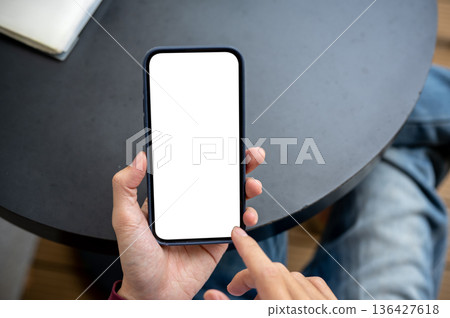 A man's hands holding white screen smartphone with finger pointing sitting at black table in a cafe. A man's hands holding white screen smartphone with finger pointing sitting at black table in a cafe. 136427618