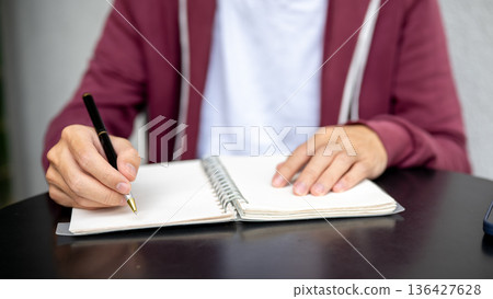 A man or student holding a ballpoint pen writing in notebook on black table in a cafe or library. A man or student holding a ballpoint pen writing in notebook on black table in a cafe or library. 136427628