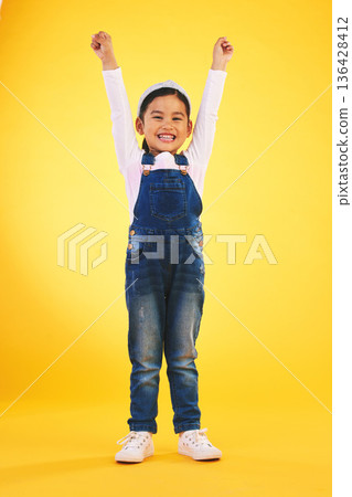Portrait, children and a winner girl on a yellow background in celebration of success or victory. Kids, goals and motivation with a young child cheering for an award or achievement in studio 136428412